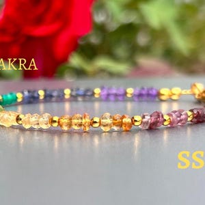 Puede incluir: Un brazalete de oro con cuentas de piedras preciosas de colores. Las cuentas están dispuestas en un patrón de arcoíris, comenzando con verde y terminando con morado. El brazalete está etiquetado como "CHAKRA" y "SSJ".