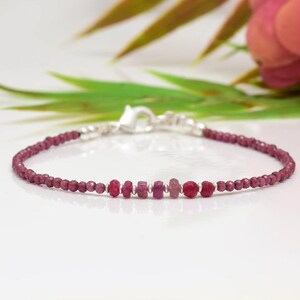 May include: A delicate bracelet featuring small, faceted, deep red beads and larger, irregularly shaped, pinkish-red gemstones. The bracelet is strung on a silver chain with a lobster clasp. The background includes green leaves and pink flowers.