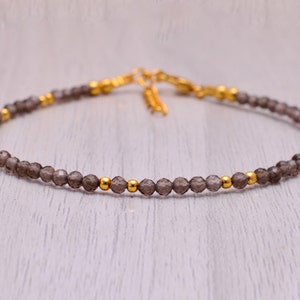 May include: A delicate bracelet made with small, faceted smoky quartz beads and gold-toned spacers. The bracelet is strung on a thin, clear elastic cord.