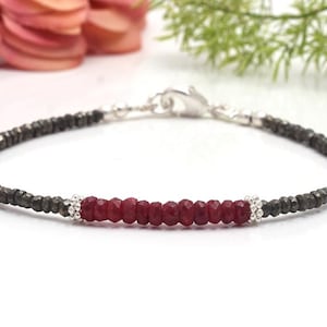 以下が含まれることがあります： 暗い灰色のパイライトビーズの2本の糸の間に、赤いルビービーズの列がセットされた、繊細なブレスレット。ブレスレットはシルバーの留め金で留められています。