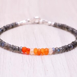 May include: A bracelet with a mix of grey, orange, and white gemstones. The bracelet has a silver clasp and is made of small, faceted beads. The central section features vibrant orange and white beads.