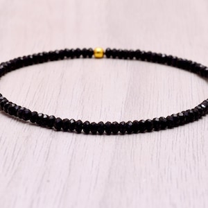 以下が含まれることがあります： 金色のアクセントビーズが付いた、繊細な黒いビーズブレスレット。ブレスレットは、小さなファセット加工された黒いビーズを繋ぎ合わせて作られています。金のビーズはブレスレットの中央に配置されています。ブレスレットは明るい色の表面に表示されています。