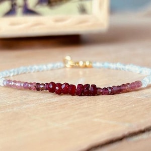 May include: A delicate bracelet with a gradient of light blue, pink, and red beads. The bracelet features a gold clasp and a row of ruby red beads in the center.