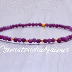 Op de afbeelding: Een delicate armband gemaakt van kleine, gefacetteerde paarse edelstenen en gouden kralen. De armband ligt op een wit houten oppervlak.