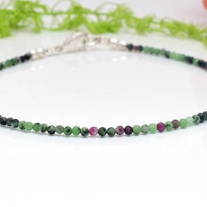 Könnte beinhalten: Ein zartes Armband aus kleinen grünen, rosa und schwarzen Edelsteinperlen. Das Armband hat einen silbernen Verschluss.