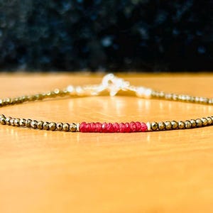 Könnte beinhalten: Ein zartes Armband aus kleinen goldfarbenen Perlen und einer Reihe roter Edelsteinperlen.