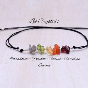 以下が含まれることがあります： 黒い紐のブレスレットに銀色のビーズと7つの小さなカラフルな宝石が並んでいます。宝石には「ラブラドライト - ペリドット - シトリン - カレリア産ガーネット」とラベルが貼られています。ブレスレットには「Leo Crystals」とラベルが貼られています。