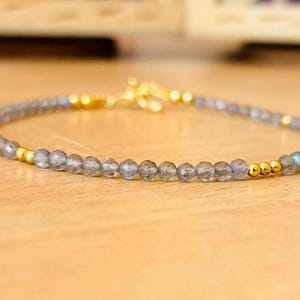 May include: A delicate bracelet made with small, faceted labradorite beads and gold accents. The bracelet is strung on a clear elastic cord.