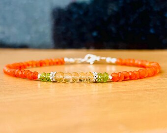 May include: A beaded bracelet featuring orange, green, and yellow gemstones. The bracelet has a silver clasp and is displayed on a wooden surface. The gemstones are small and round, with a vibrant color palette.