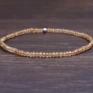 May include: A delicate bracelet composed of small, faceted, amber-colored beads. The beads are strung together, forming a circular shape. A small, silver-colored clasp secures the bracelet. The jewelry is displayed on a dark brown wooden surface.