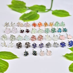 May include: Assortment of 24 pairs of silver stud earrings with various semi-precious gemstones. The stones are raw and have a natural, rough texture. The earrings are arranged in rows on a white surface with green leaves.