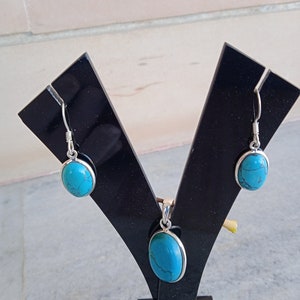 May include: A turquoise stone pendant and matching earrings set on a black display stand. The oval-shaped stones are set in silver.