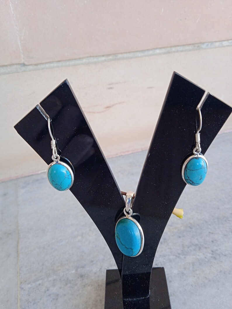 May include: A turquoise stone pendant and matching earrings set on a black display stand. The oval-shaped stones are set in silver.