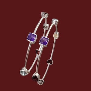 May include: Three silver bangles with various gemstones. The bangles feature square purple amethyst stones, round clear stones, and heart-shaped red stones. The background is a solid maroon color.