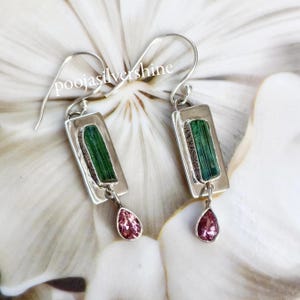 May include: Silver earrings featuring green tourmaline stones and pink teardrop-shaped stones. The earrings are rectangular in shape and have a hammered texture.