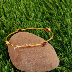 Pode incluir: Uma pulseira dourada com quatro pedras preciosas ovais de cor vermelha escura. A pulseira está sobre uma pedra castanha contra um fundo de relva verde. O texto "poojasilvershine" é visível na pedra.