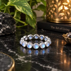 May include: A circular ring featuring a continuous row of round, bezel-set moonstones. Each stone displays a translucent, milky white appearance with a distinct blue adularescence, set within a polished silver-toned metal band.