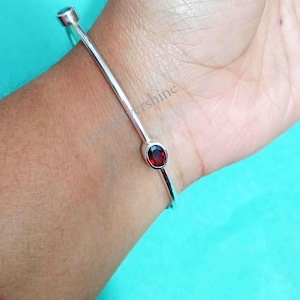 Op de afbeelding: Een zilverkleurige armband met een lineair ontwerp, met twee ovale, dieprode edelstenen. De armband wordt om een pols gedragen en toont zijn minimalistische stijl en het contrast tussen het metaal en de edelstenen.