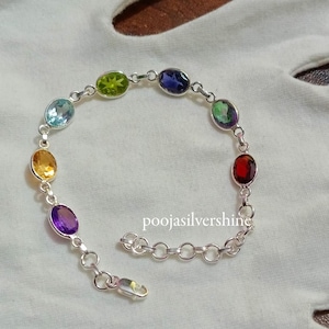 Puede incluir: Una pulsera de plata con siete piedras preciosas ovaladas en colores del arcoíris. La pulsera tiene un cierre de langosta. El texto "poojasilvershine" es visible en la pulsera.