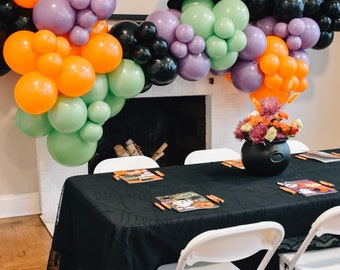 May include: A black tablecloth with a spiderweb pattern covers a table with white folding chairs. A black cauldron sits on the table with a bouquet of purple and orange flowers. A garland of orange, purple, green, and black balloons hangs above the table. The sign above the garland reads "hello pumpkin".