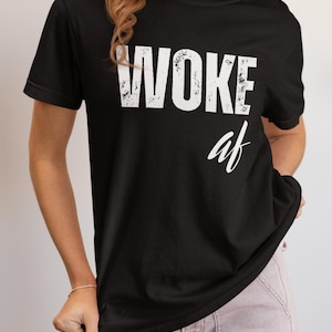 May include: Black crew neck t-shirt with the words "WOKE af" printed in white. The shirt is worn by a person wearing light pink pants.