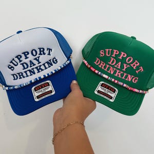 May include: Two trucker hats with the text "Support Day Drinking" embroidered on the front. The hats are blue and green with white mesh panels. Both hats have a colorful beaded trim.