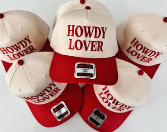 Peut inclure: Cinq casquettes de baseball blanches avec visières rouges. Chaque casquette a le texte "Howdy Lover" brodé en rouge sur le devant.