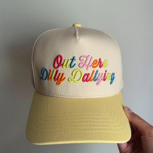 May include: A cream-colored baseball cap with a yellow brim and a yellow button on top. The front features the embroidered text Out Here Dilly Dallying in multi-colored cursive lettering, including shades of pink, blue, orange, and lime green.