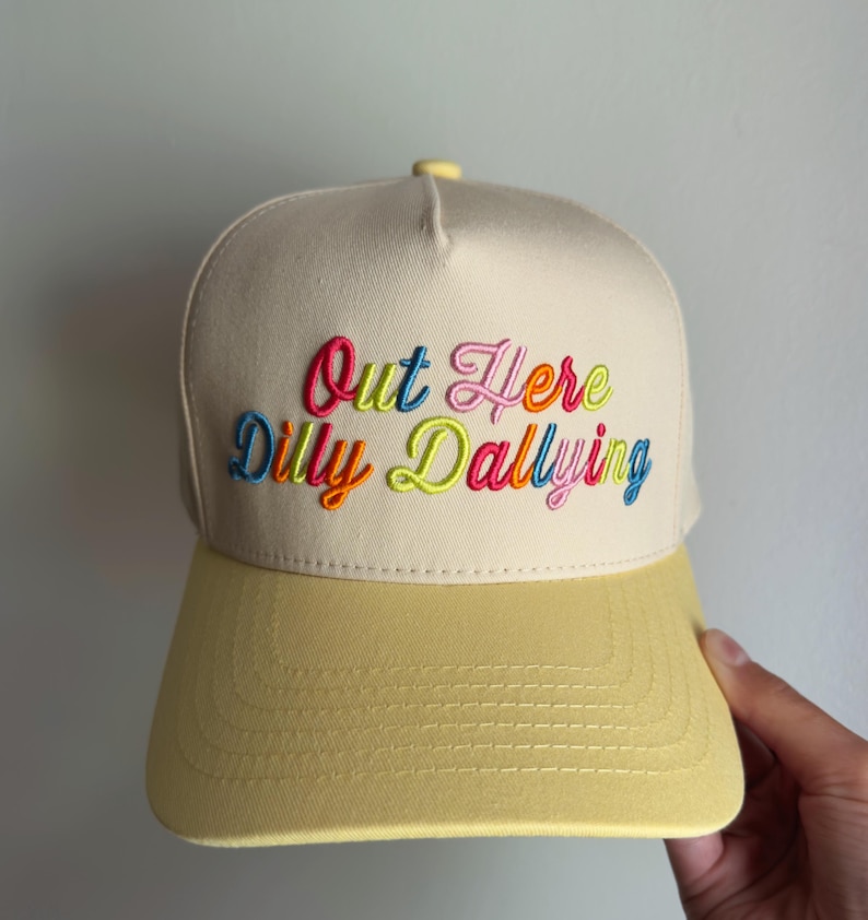 May include: A cream-colored baseball cap with a yellow brim and a yellow button on top. The front features the embroidered text Out Here Dilly Dallying in multi-colored cursive lettering, including shades of pink, blue, orange, and lime green.