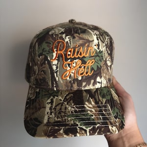 May include: A camouflage baseball cap with the embroidered text "Raisin Hell" in orange script. The hat features a curved brim and a structured crown, with a pattern of green, brown, and tan foliage.