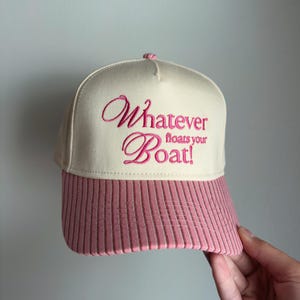 May include: A cream-colored baseball cap with a pink corduroy brim. The front panel features the embroidered pink text "Whatever floats your Boat!" in a mix of cursive and block lettering. The cap has a structured crown and a top button.
