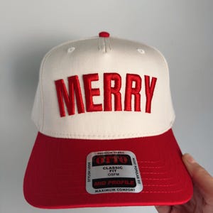 May include: A white and red baseball cap with the word "MERRY" embroidered in red. The cap has a red brim and a button on top. The tag reads "OTTO CLASSIC FIT OSFM MID PROFILE MAXIMUM COMFORT".