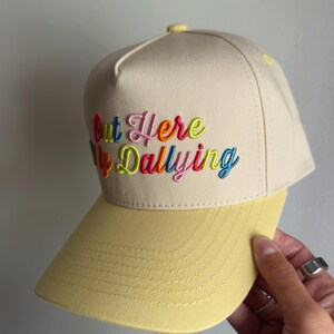 May include: A beige baseball cap with a yellow brim and a structured crown. The front features multi-colored embroidered text that reads Out Here Dallying, with each letter stitched in vibrant shades of pink, orange, blue, and lime green.