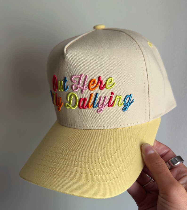 May include: A beige baseball cap with a yellow brim and a structured crown. The front features multi-colored embroidered text that reads Out Here Dallying, with each letter stitched in vibrant shades of pink, orange, blue, and lime green.