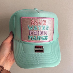 May include: A turquoise trucker hat with a pink, white, and blue embroidered patch that says "Save Water Drink Margs". The hat also has a black label that says "Otto World Famous Foam Front Otto Trucker".