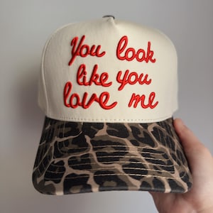 May include: A cream-colored baseball cap with the embroidered phrase "You look like you love me" in red script. The brim features a brown and black animal print pattern. A stylish and unique accessory.
