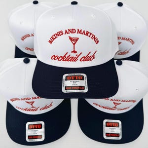 May include: Five white and navy blue baseball caps with the text "Bikinis and Martinis cocktail club" in red and white lettering. Each cap has a red martini glass graphic and a black and white label with the text "OTTO" in white.
