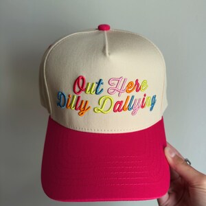 May include: A beige baseball cap with a bright pink brim and a matching pink button on top. The front panel features the embroidered text "Out Here Dilly Dallying" in a multi-colored script font, using shades of blue, pink, orange, and yellow.