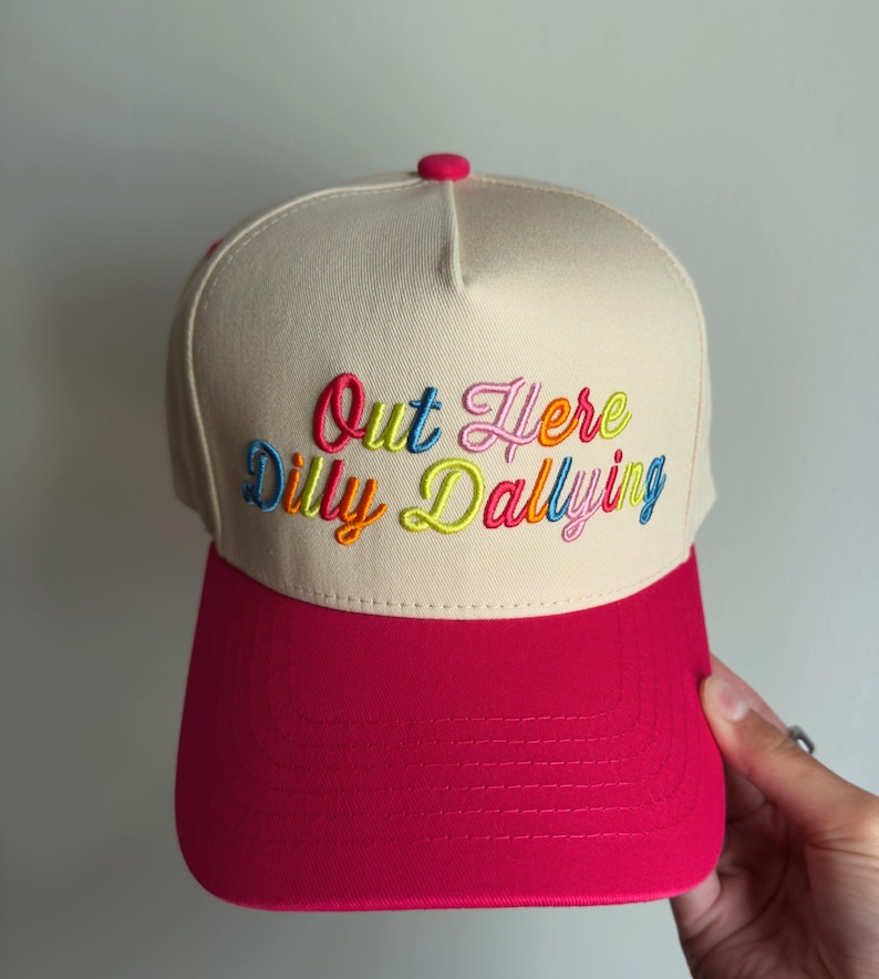 May include: A beige baseball cap with a bright pink brim and a matching pink button on top. The front panel features the embroidered text "Out Here Dilly Dallying" in a multi-colored script font, using shades of blue, pink, orange, and yellow.