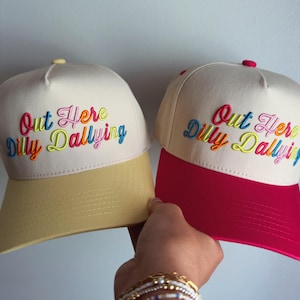 May include: Two baseball caps with off-white crowns and embroidered text that reads Out Here Dilly Dallying in multi-colored script. One cap features a light yellow brim and button, while the other has a vibrant pink brim and matching top button.