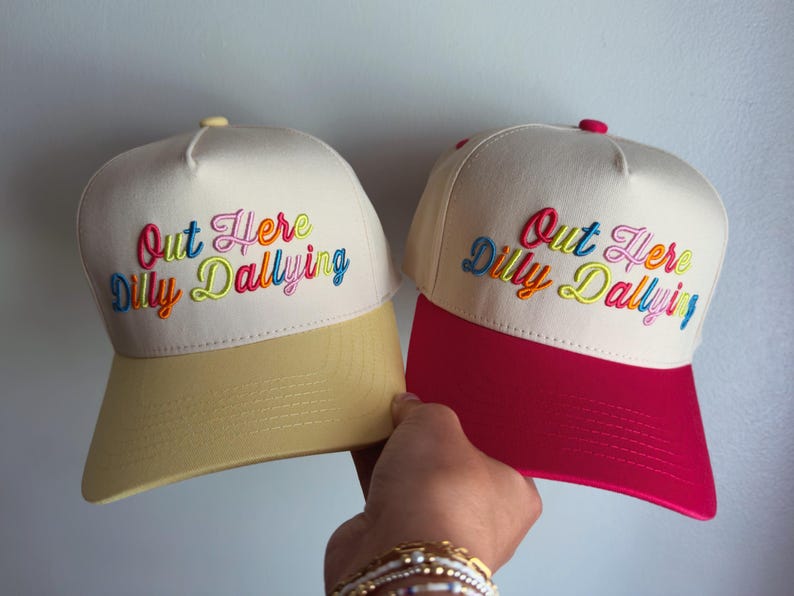 May include: Two baseball caps with off-white crowns and embroidered text that reads Out Here Dilly Dallying in multi-colored script. One cap features a light yellow brim and button, while the other has a vibrant pink brim and matching top button.