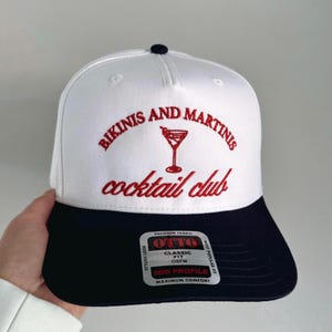May include: White and navy blue baseball cap with red embroidery that reads "Bikinis and Martinis cocktail club". The cap has a logo with a martini glass and the words "Otto Classic Fit OSFM Mid Profile Maximum Comfort" on a white tag.