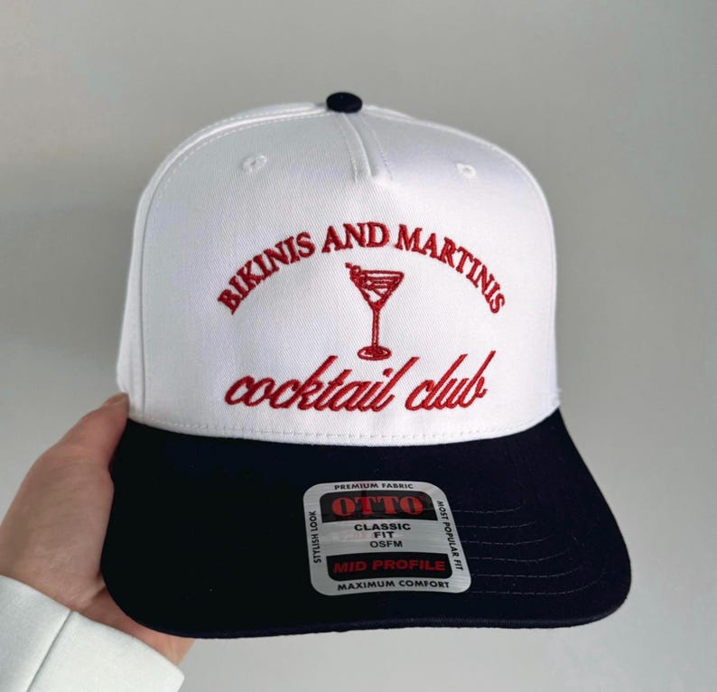 May include: White and navy blue baseball cap with red embroidery that reads "Bikinis and Martinis cocktail club". The cap has a logo with a martini glass and the words "Otto Classic Fit OSFM Mid Profile Maximum Comfort" on a white tag.