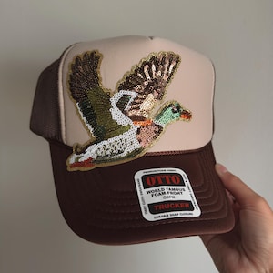 May include: A beige and brown trucker hat featuring a large, sequined mallard duck patch on the front panel. The hat includes a white sticker with the text OTTO WORLD FAMOUS FOAM FRONT OSFM TRUCKER, indicating premium foam fabric and a snap closure.