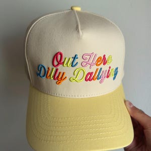 May include: A cream-colored baseball cap with a yellow brim and a yellow button on top. The front features multi-colored embroidered text that reads Out Here Dilly Dallying in a cursive font with pink, blue, orange, and lime green thread.