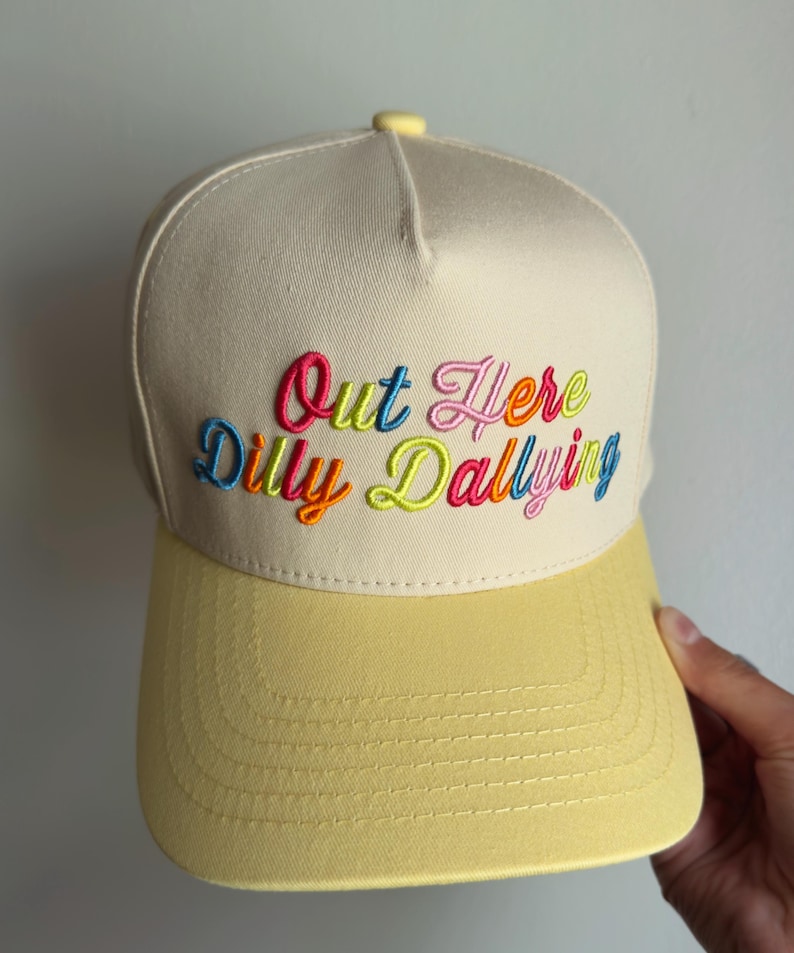 May include: A cream-colored baseball cap with a yellow brim and a yellow button on top. The front features multi-colored embroidered text that reads Out Here Dilly Dallying in a cursive font with pink, blue, orange, and lime green thread.
