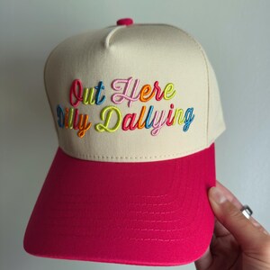 May include: A beige baseball cap with a bright pink brim and a small pink button on top. The front features multi-colored embroidered text that reads Out Here Dilly Dallying in a cursive font with letters in shades of blue, pink, orange, and green.
