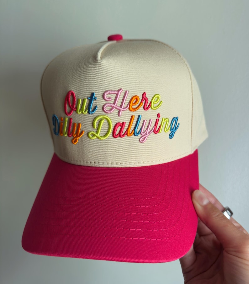 May include: A beige baseball cap with a bright pink brim and a small pink button on top. The front features multi-colored embroidered text that reads Out Here Dilly Dallying in a cursive font with letters in shades of blue, pink, orange, and green.