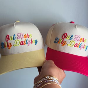 May include: Two cream-colored baseball caps with embroidered text that reads Out Here Dilly Dallying in multi-colored script. One cap features a pale yellow brim, while the other cap features a vibrant pink brim. Both caps have a structured crown.