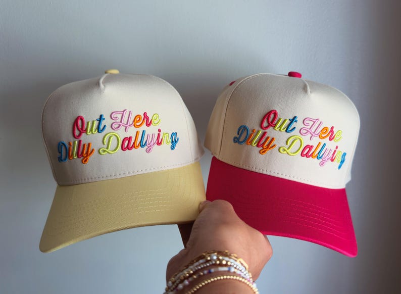 May include: Two cream-colored baseball caps with embroidered text that reads Out Here Dilly Dallying in multi-colored script. One cap features a pale yellow brim, while the other cap features a vibrant pink brim. Both caps have a structured crown.